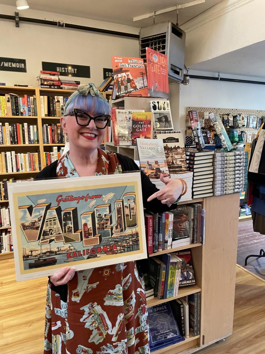 Alibi Bookshop owner Karen Finlay poses with old-fashioned Vallejo Postcard poster in front of the Local Authors section. Photo courtesy of Karen Finlay. 