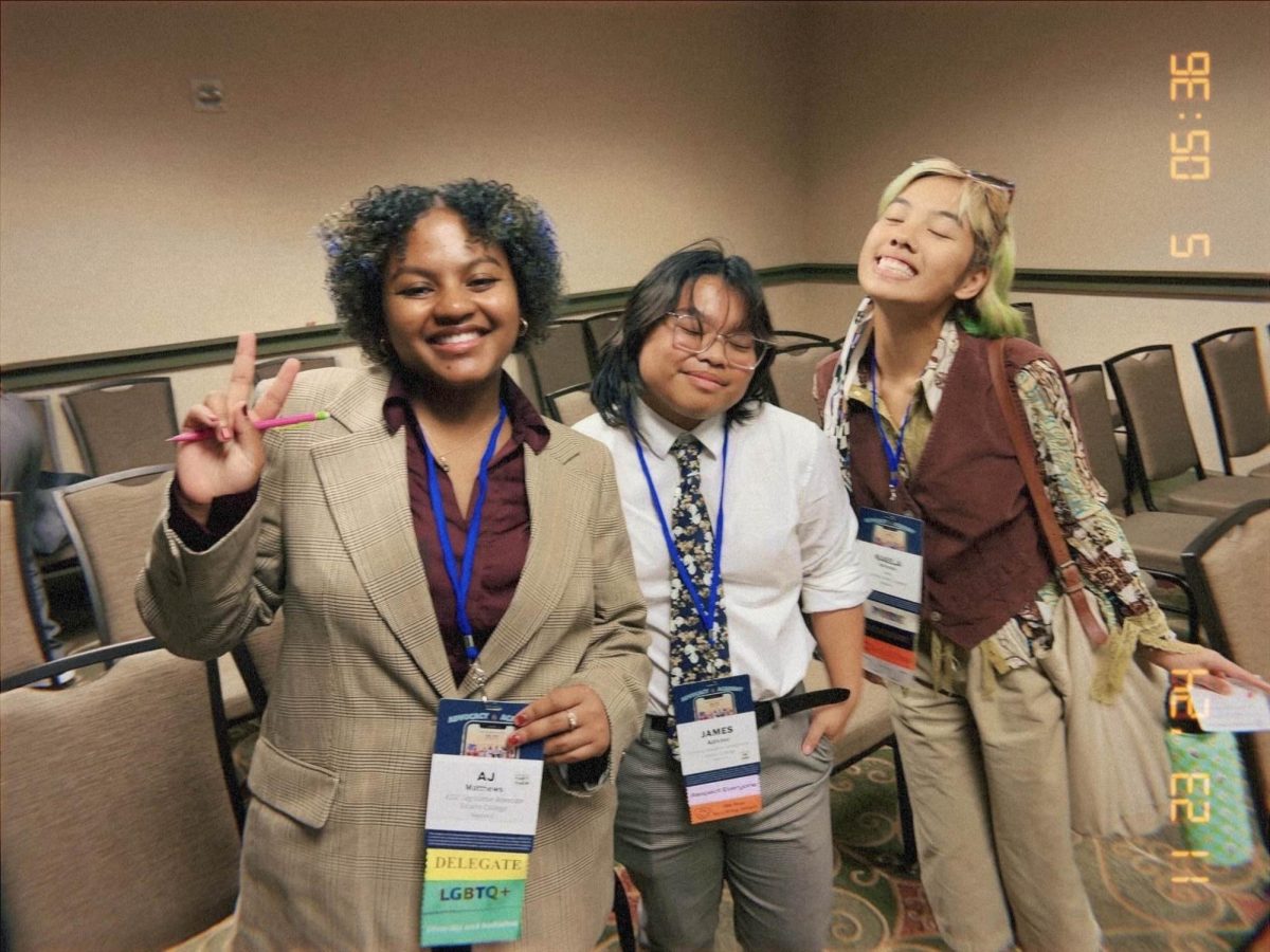 The image features 3 people, The closet one to the camera is AJ Matthews in a blazer. They have a tan blazer and purple undershirt. Their also smiling with a wide grin. Next to them is two of their friends James Adivino and Kaela Sarsoza. James is wearing a white collared shirt, khaki pants, and a tie while Kaela is wearing a red shirt and khaki pants.